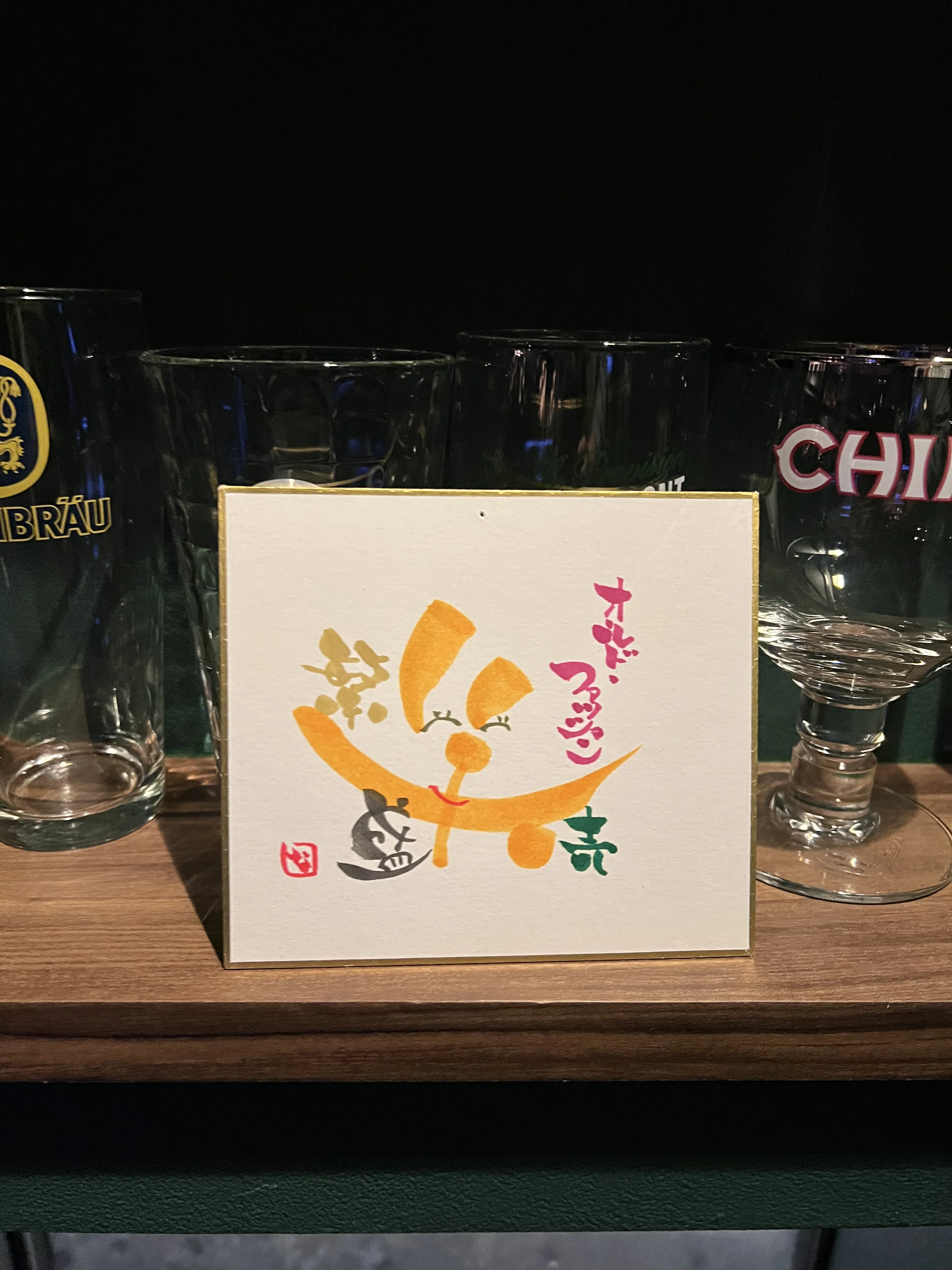 夜の入口近くに置かれた小さな看板と店内へ続く灯り、奥に並ぶグラス